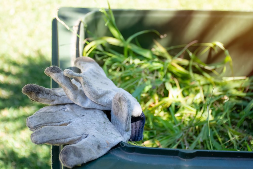 Green bin container filled with garden waste. Dirty gardening gloves. Spring clean up in the garden. Recycling garbage for a better environment.